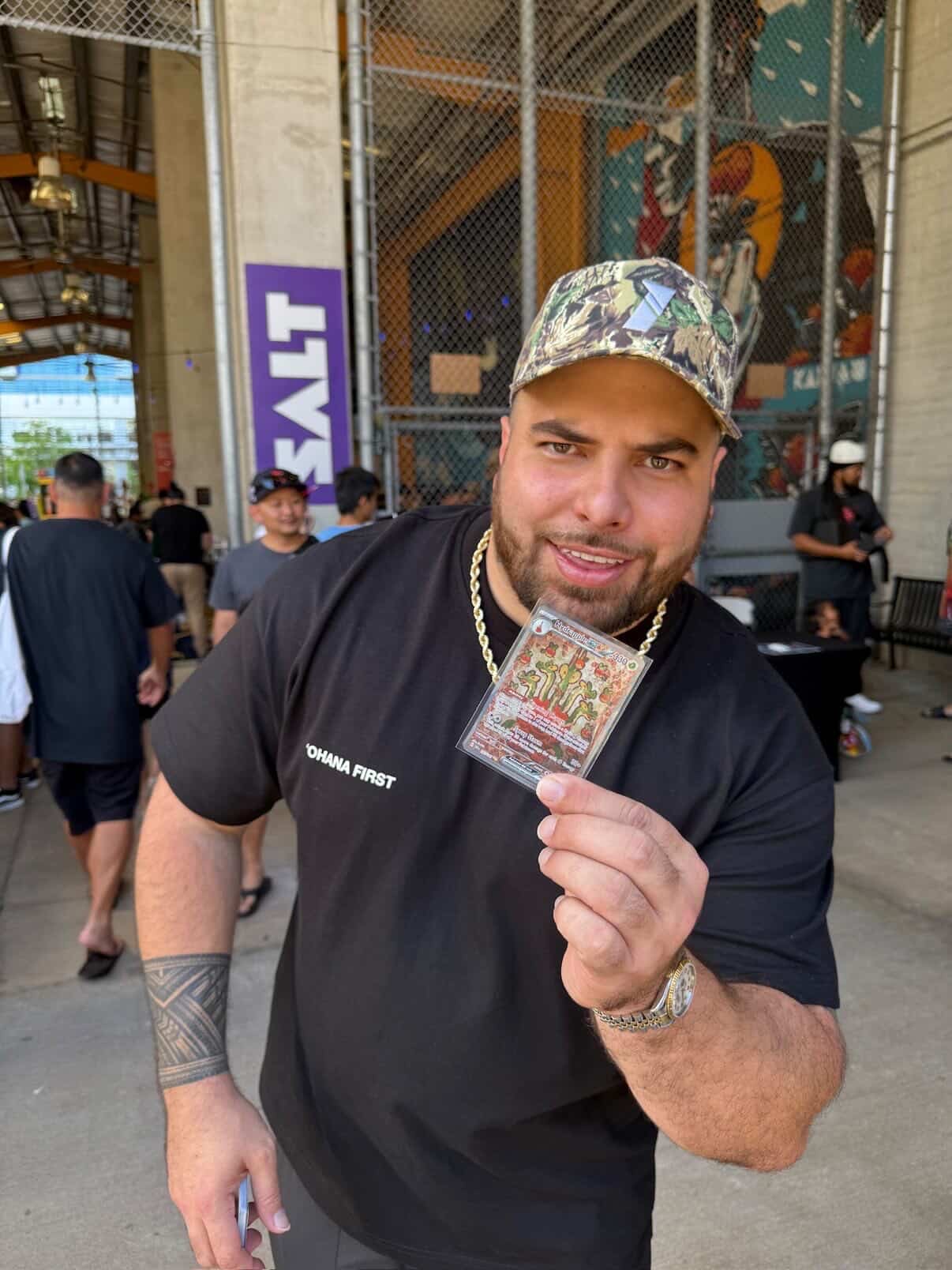 Keep It Aloha Card Show — attendee in OHANA FIRST tee holding up a Pokemon card pull at the SALT Kakaako entrance